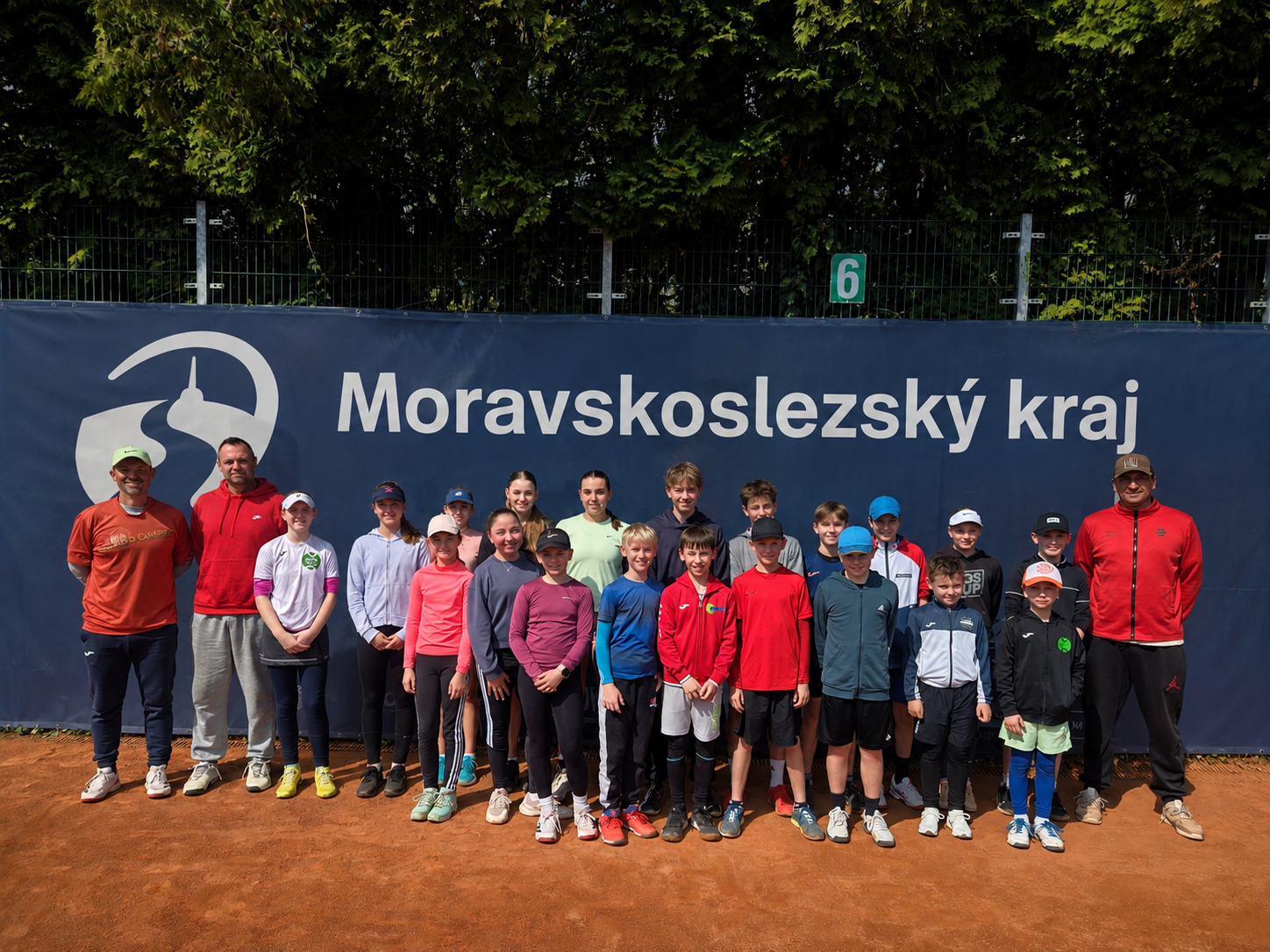 Skupinová fotka dětí a trenérů na tenisovém kurtu před velkým bannerem s nápisem.